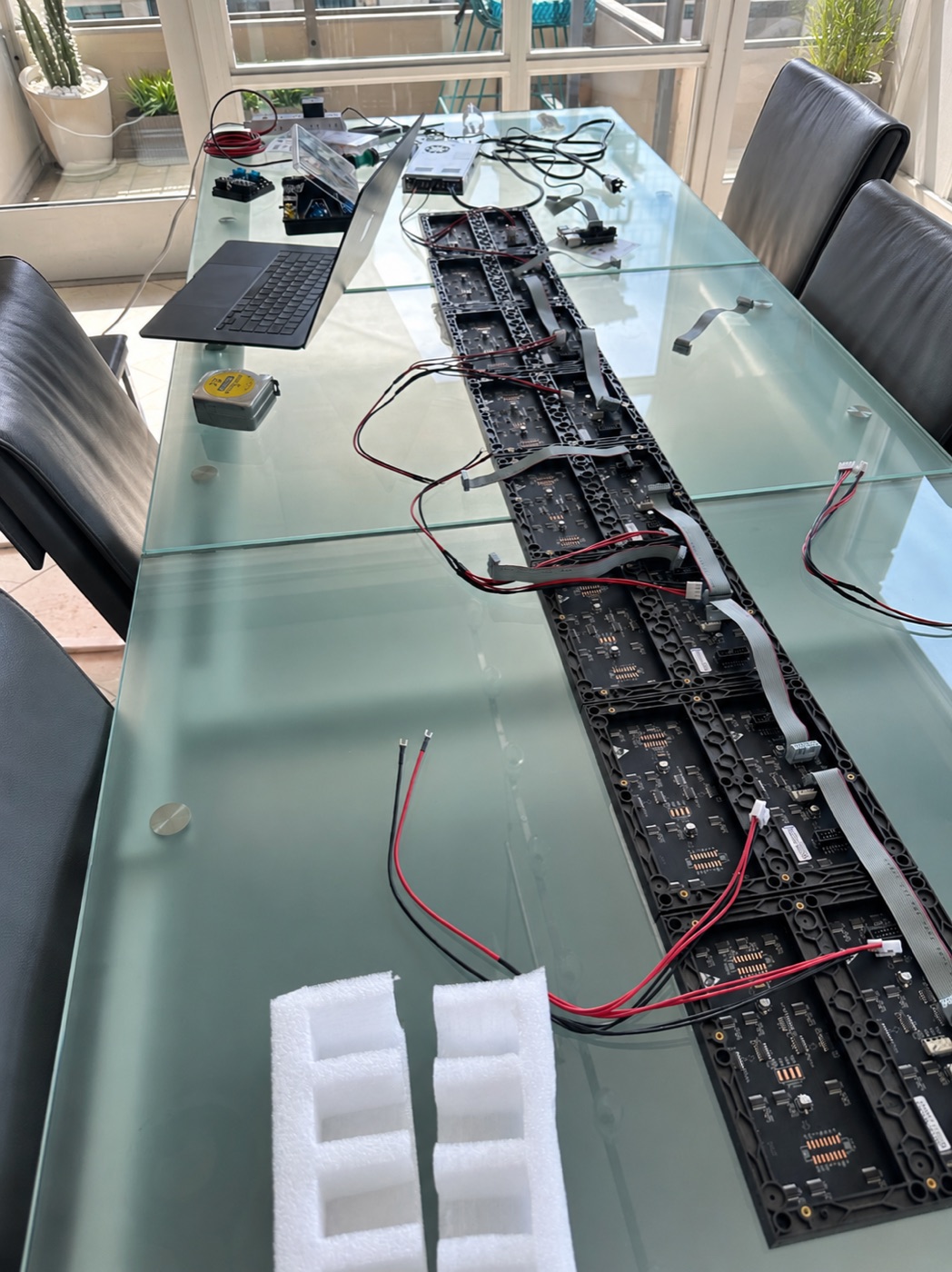 The seven panels laid face down in a row on a glass dining table, ribbon cables and red and black power leads between them, with the power supply, fuse block and a laptop at the far end