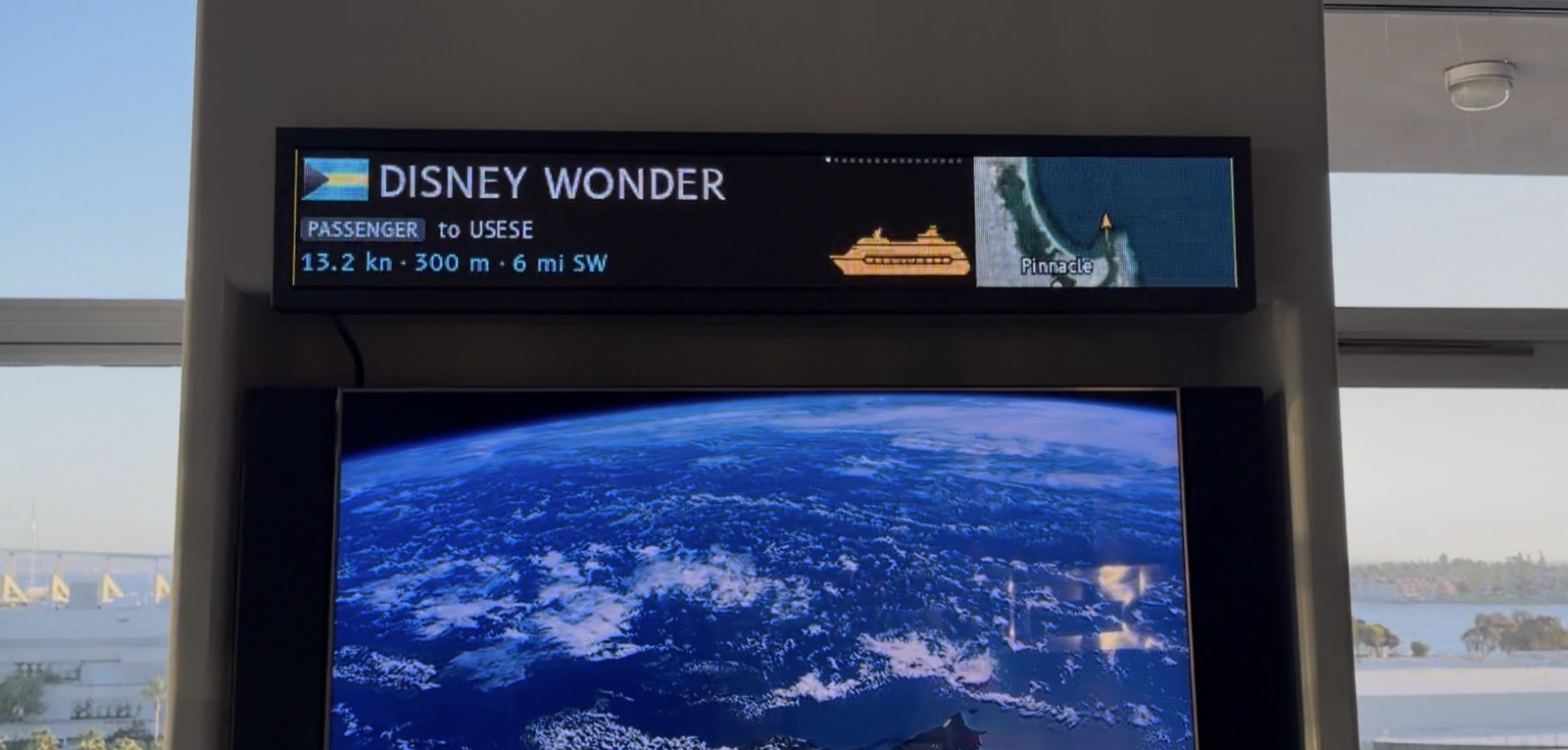 The LumiBridge display mounted on a pillar above a TV, in front of windows onto San Diego Bay, showing the cruise ship Disney Wonder with its speed, length and distance beside a map of the bay